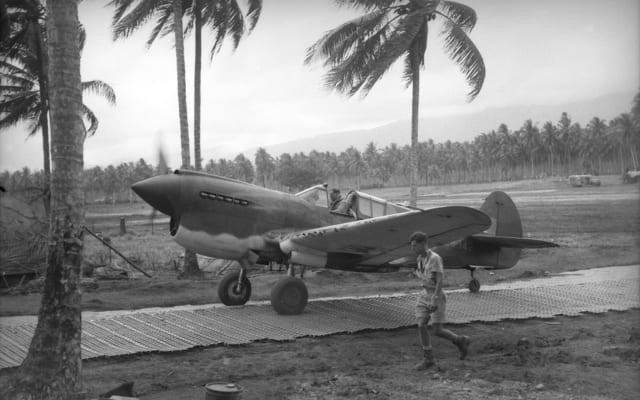 A P-40 Kittyhawk returns from an operational flight and taxis up the metal runway to the dispersabl bays set among coconut trees.